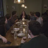 Friends round a long restaurant table laughing with the birthday woman, a cake with one candle in front of her and one man sitting a little back