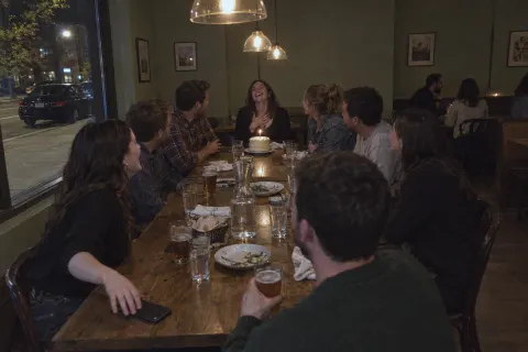 Friends round a long restaurant table laughing with the birthday woman, a cake with one candle in front of her and one man sitting a little back