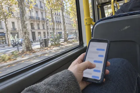 A hand holds a phone with a message thread on a bus, autumn trees passing outside the window