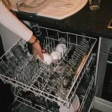 A hand puts a cup into the top rack of an open dishwasher, wine glasses waiting on the counter