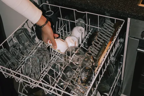 A hand puts a cup into the top rack of an open dishwasher, wine glasses waiting on the counter