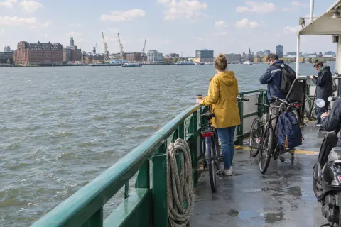 A woman in a yellow raincoat stands with her bike at the rail of a city ferry, looking across the water