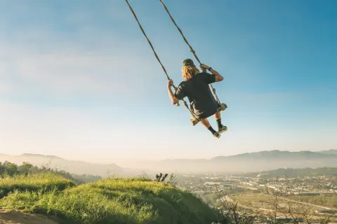 A grown-up on a rope swing, swung far out over a green hilltop against a clear blue sky