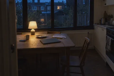 A kitchen table by a big window at the end of the day, one lamp on and a closed laptop