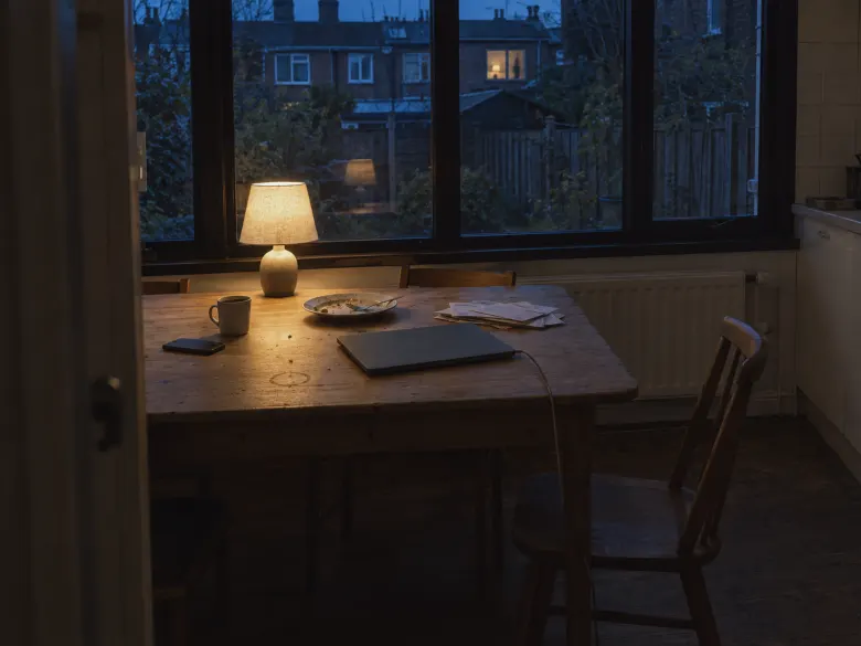 A kitchen table by a big window at the end of the day, one lamp on and a closed laptop