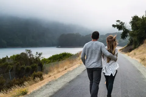 A couple seen from behind walking down a quiet road by a lake, his arm around her shoulders, mist on the hills