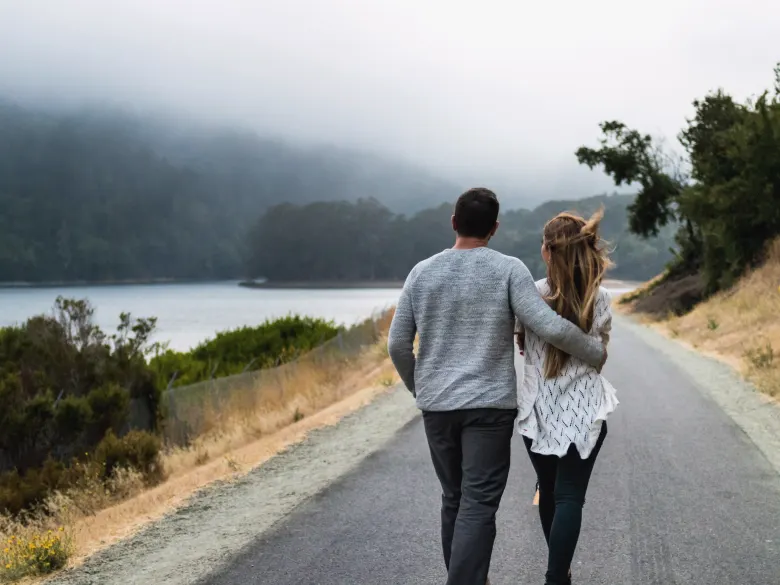 A couple seen from behind walking down a quiet road by a lake, his arm around her shoulders, mist on the hills