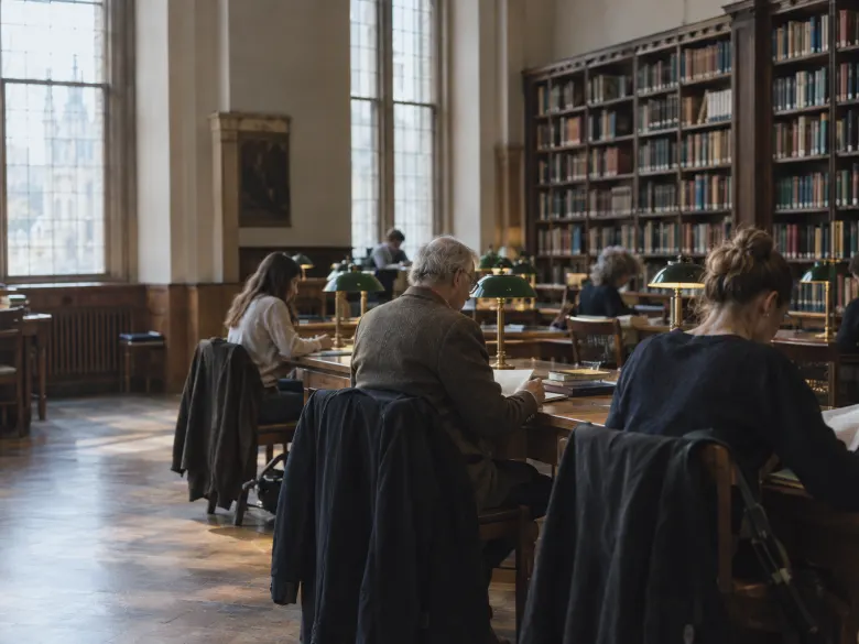 People reading at long wooden tables with green lamps in an old library reading room