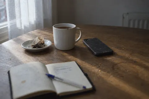 A small open notebook and a pen, a mug of tea and a phone on a wooden table by the window