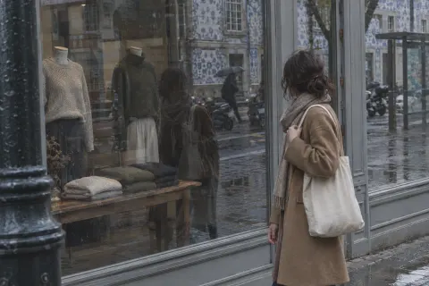 A woman on a rainy shopping street catches her own reflection in a shop window