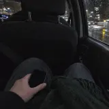 A hand holding a phone on the back seat of a car at night, city lights streaking past in the rain