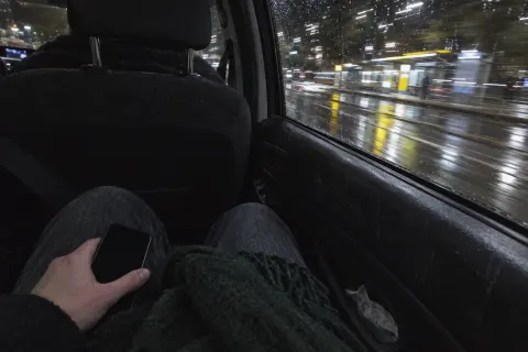 A hand holding a phone on the back seat of a car at night, city lights streaking past in the rain