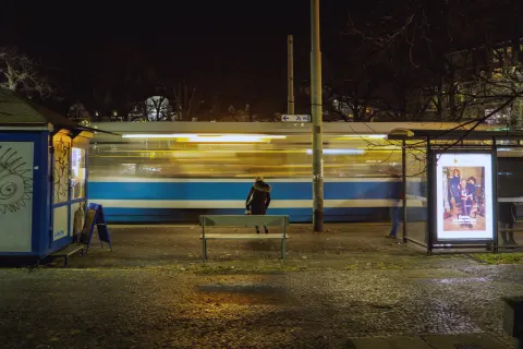 Someone waiting at a tram stop at night, seen from behind, as a tram streaks past