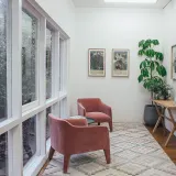 Two pink armchairs turned towards each other beside a wall of tall windows in a bright white room with a large plant.